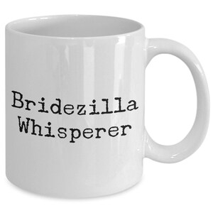 May include: White ceramic coffee mug with black text that reads "Bridezilla Whisperer".