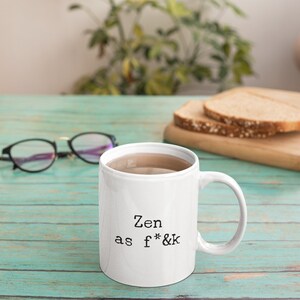 May include: White ceramic mug with the text "Zen as f*&k" in black. The mug is filled with a light brown liquid. The mug is on a teal wooden surface, with eyeglasses and a cutting board with bread slices in the background.