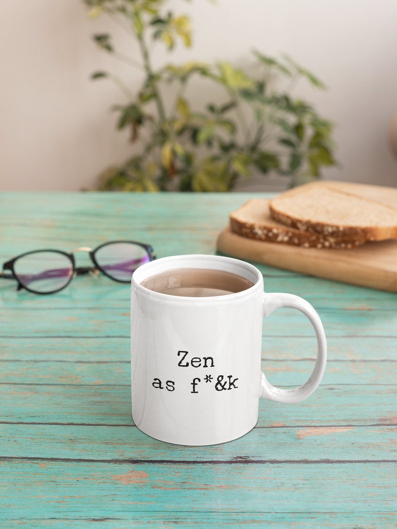 May include: White ceramic mug with the text "Zen as f*&k" in black. The mug is filled with a light brown liquid. The mug is on a teal wooden surface, with eyeglasses and a cutting board with bread slices in the background.