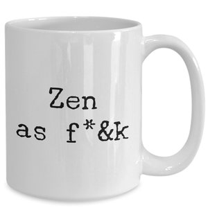 May include: White ceramic mug with a curved handle. The mug features the phrase "Zen as f*&k" printed in black, bold, typewriter-style font. The mug is designed for hot beverages.