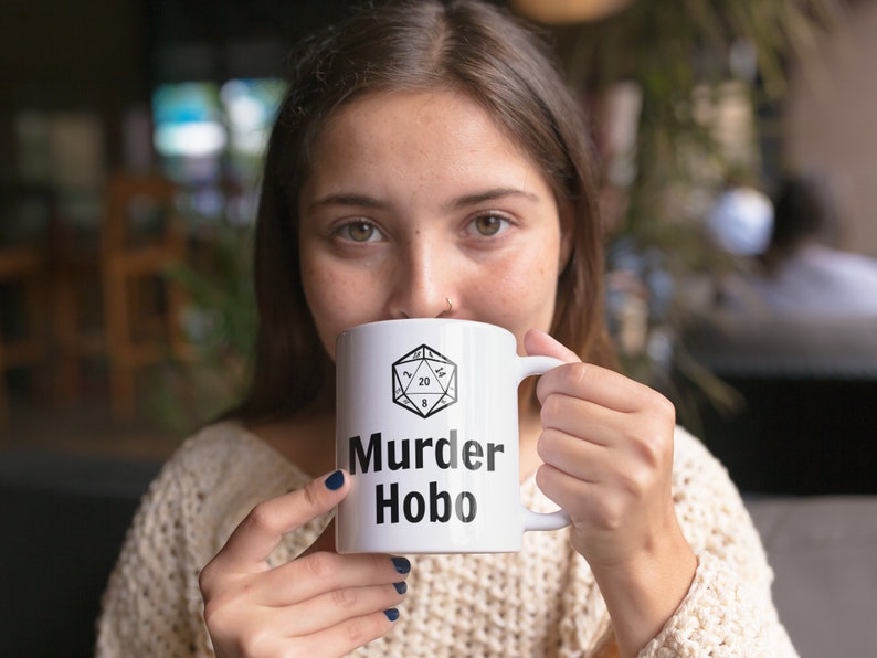 K&ouml;nnte beinhalten: Eine wei&szlig;e Keramiktasse mit einer schwarz-wei&szlig;en Illustration eines zwanzigseitigen W&uuml;rfels und dem Text "Murder Hobo".