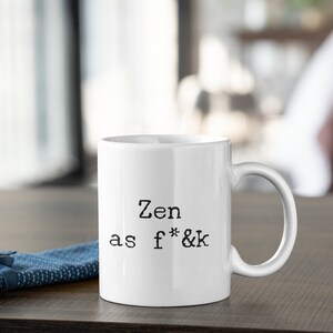 May include: White ceramic mug with the text "Zen as f*&k" in black, sans-serif font. The mug has a curved handle and sits on a dark wooden surface. A blue fabric is visible on the left.