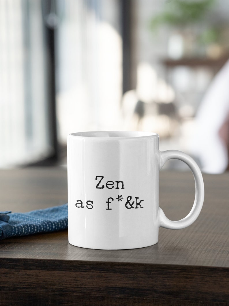 May include: White ceramic mug with the text "Zen as f*&k" in black, sans-serif font. The mug has a curved handle and sits on a dark wooden surface. A blue fabric is visible on the left.