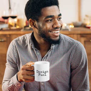 May include: A person holding a white ceramic mug with the text "Design Fuel" in black lettering.