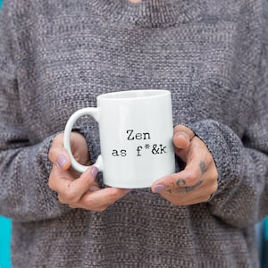 May include: White ceramic mug with the text "Zen as f*&k" in black, held by a person wearing a gray sweater. The mug has a standard handle and a simple, clean design, suitable for coffee or tea.