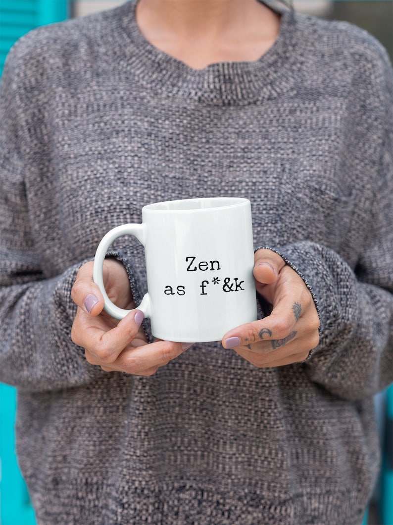 May include: White ceramic mug with the text "Zen as f*&k" in black, held by a person wearing a gray sweater. The mug has a standard handle and a simple, clean design, suitable for coffee or tea.