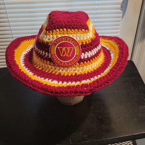 May include: A crocheted cowboy hat featuring horizontal stripes in burgundy, gold, and white yarn. The front of the hat displays a circular patch with the Washington Commanders logo, featuring a yellow W on a burgundy background with gold text.