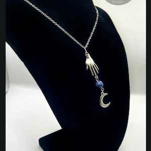 May include: A silver-toned necklace featuring a delicate chain, a pendant shaped like a human hand, a small round blue speckled bead, and a silver crescent moon charm hanging vertically from the hand, creating a layered pendant design.