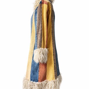 May include: A long coat featuring vertical panels of blue, yellow, and rust-colored denim fabric. The garment is accented with cream-colored faux fur trim along the collar, the sleeve cuffs, and the bottom hemline of the coat.
