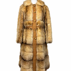 May include: A long, full-length coat featuring a combination of tan, brown, and golden-toned faux fur panels. The garment includes a central vertical suede-like strip down the front, secured by five round, light-colored wooden buttons.