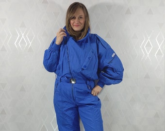 May include: A bright blue ski jumpsuit with a zipper down the front and a belt. The jumpsuit has a high collar and a logo on the sleeve.