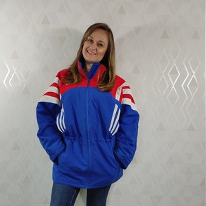 Op de afbeelding: Een blauwe en rode vintage Adidas jas met witte strepen en een trekkoord in de taille. De jas wordt gedragen door een persoon met lang bruin haar en staat op een houten vloer.