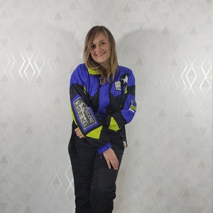 Pode incluir: Uma mulher vestindo uma jaqueta de esqui azul, preta e amarela neon com o texto "The Scene Is Back" na manga esquerda. Ela tamb&eacute;m est&aacute; vestindo cal&ccedil;as pretas e t&ecirc;nis brancos.