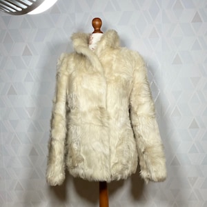 Könnte beinhalten: Eine weiße Kunstfelljacke mit Kragen und langen Ärmeln. Die Jacke wird auf einem Holz-Mannequin präsentiert.