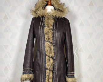 Può includere: Un lungo cappotto di pelliccia sintetica marrone con cappuccio e bordatura in pelliccia. Il cappotto ha una chiusura a zip e una chiusura a bottone. Il cappotto è foderato con un tessuto marrone.