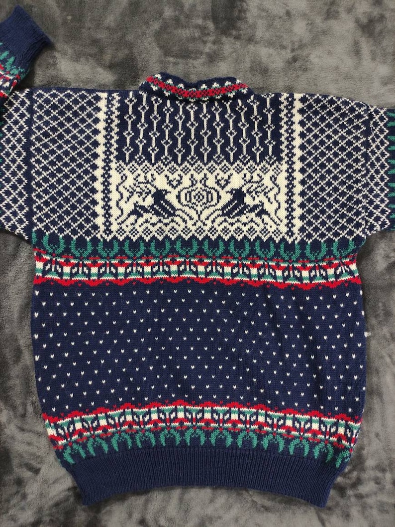 Puede incluir: Un su&eacute;ter de punto azul con un dise&ntilde;o estampado en blanco, rojo y verde. El su&eacute;ter presenta un dise&ntilde;o escandinavo tradicional con renos y patrones geom&eacute;tricos.