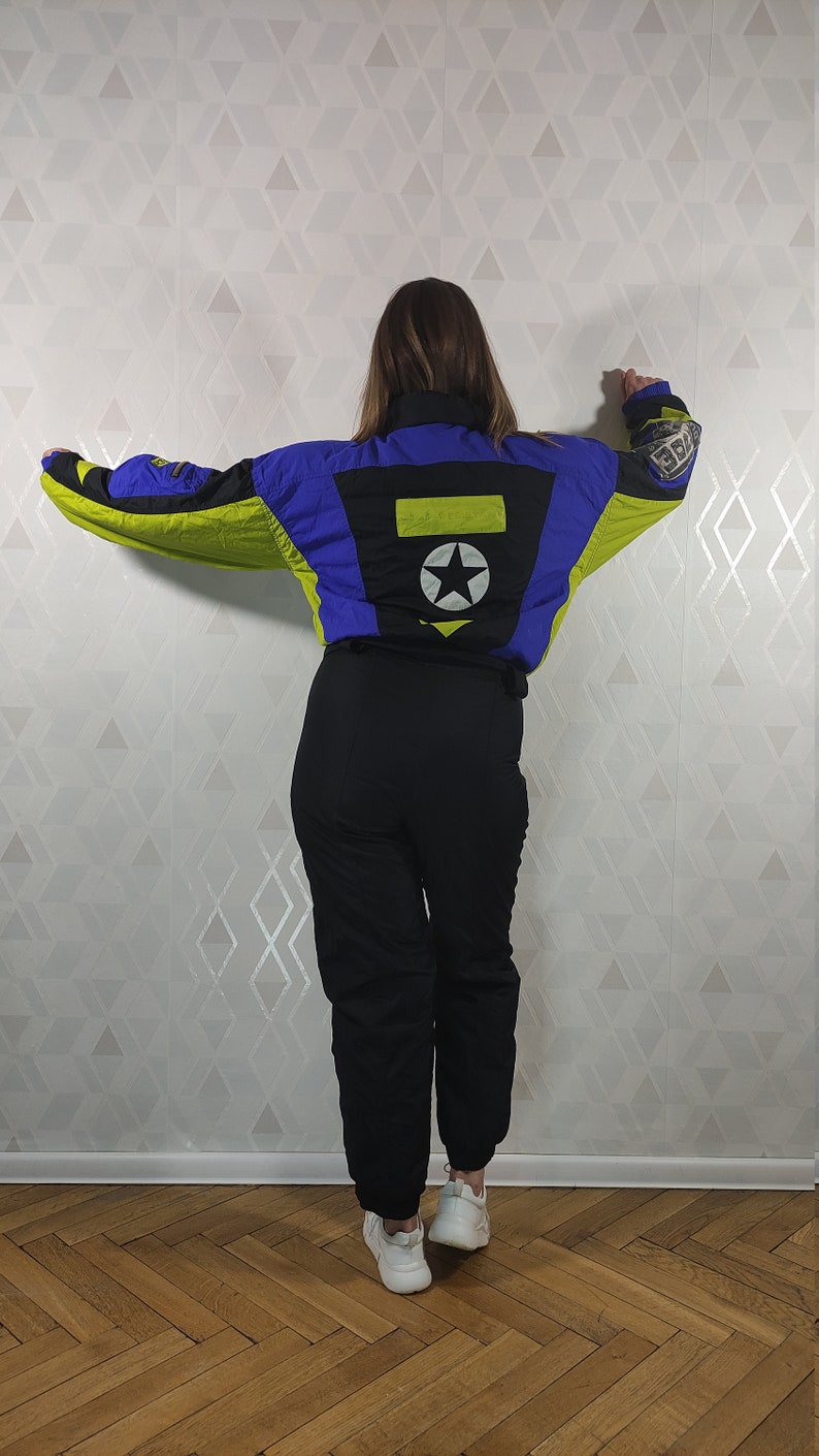 Pode incluir: Um macac&atilde;o de esqui preto e azul com detalhes amarelos. O macac&atilde;o tem uma estrela e um ret&acirc;ngulo nas costas. A pessoa est&aacute; usando t&ecirc;nis brancos.