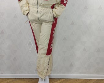 May include: A cream and red one-piece ski suit featuring a front zipper, elasticized waist, and side pockets. The chest displays a circular graphic with white line art, while the sleeves and pant legs feature the text "THALASSA" in white.