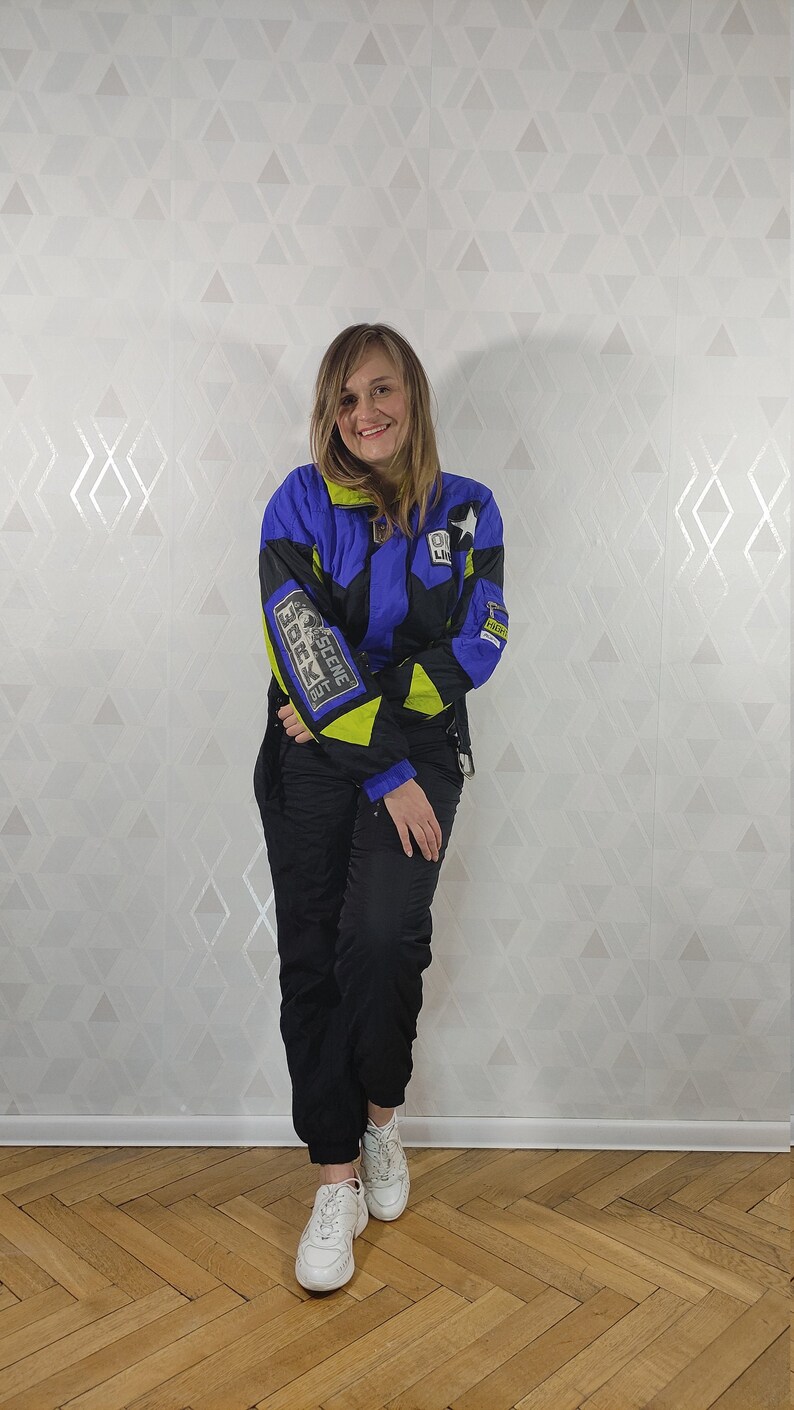 Pode incluir: Uma mulher vestindo uma jaqueta de esqui azul, preta e amarela neon com o texto "The Scene Is Back" na manga esquerda. Ela tamb&eacute;m est&aacute; vestindo cal&ccedil;as pretas e t&ecirc;nis brancos.
