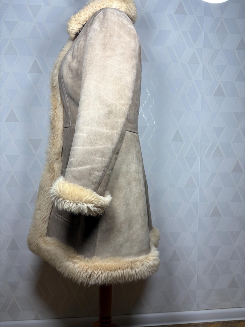 Puede incluir: Un abrigo de ante marr&oacute;n con forro y ribete de borreguillo color crema. El abrigo presenta un dise&ntilde;o cl&aacute;sico con cuello con solapas, mangas largas y un dobladillo hasta la rodilla. El abrigo se exhibe en un soporte de madera.