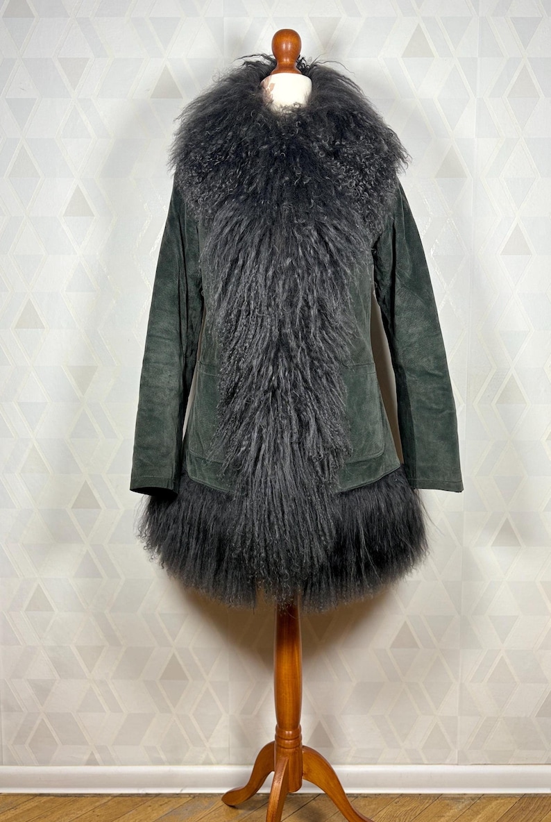 May include: A long, dark green suede coat with a large, fluffy black fur collar and trim. The coat has a long, fitted silhouette and a single pocket on the left side.