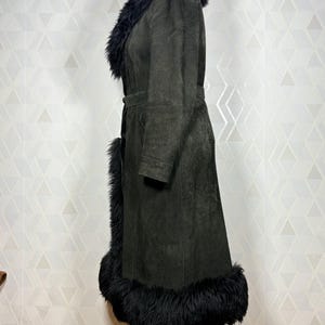 Puede incluir: Un abrigo de ante verde oscuro con cuello, pu&ntilde;os y dobladillo de piel negra. El abrigo tiene una cintura con cintur&oacute;n y un dise&ntilde;o largo y fluido. El abrigo se exhibe en un maniqu&iacute; de madera.