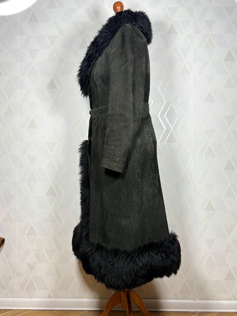 Puede incluir: Un abrigo de ante verde oscuro con cuello, pu&ntilde;os y dobladillo de piel negra. El abrigo tiene una cintura con cintur&oacute;n y un dise&ntilde;o largo y fluido. El abrigo se exhibe en un maniqu&iacute; de madera.