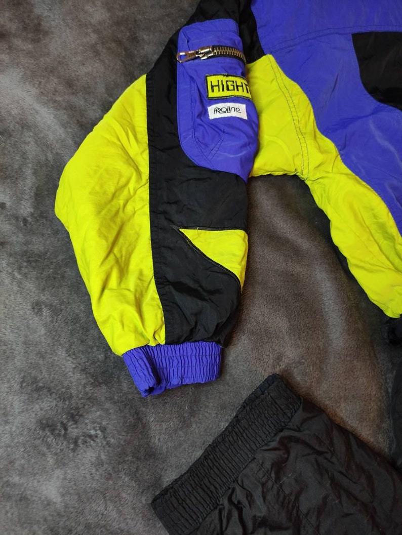 Pode incluir: Uma jaqueta de esqui preta, azul e amarela com um z&iacute;per preto e um remendo amarelo que diz "HIGHTRAILINE". A jaqueta &eacute; combinada com cal&ccedil;as de esqui pretas.