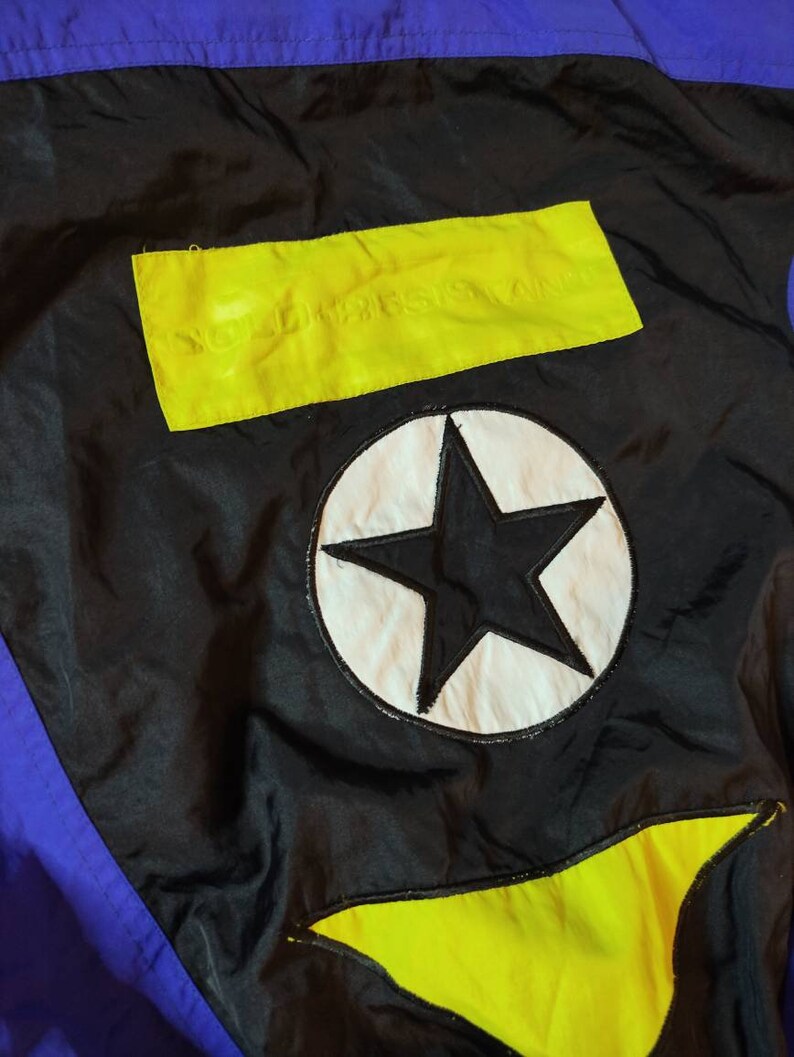 Pode incluir: Uma jaqueta de nylon preta com um design de blocos de cores amarelo e azul. A jaqueta tem uma grande estrela branca com contorno preto nas costas. O texto "NORTHWEST AIRLINES" est&aacute; impresso em um ret&acirc;ngulo amarelo nas costas.