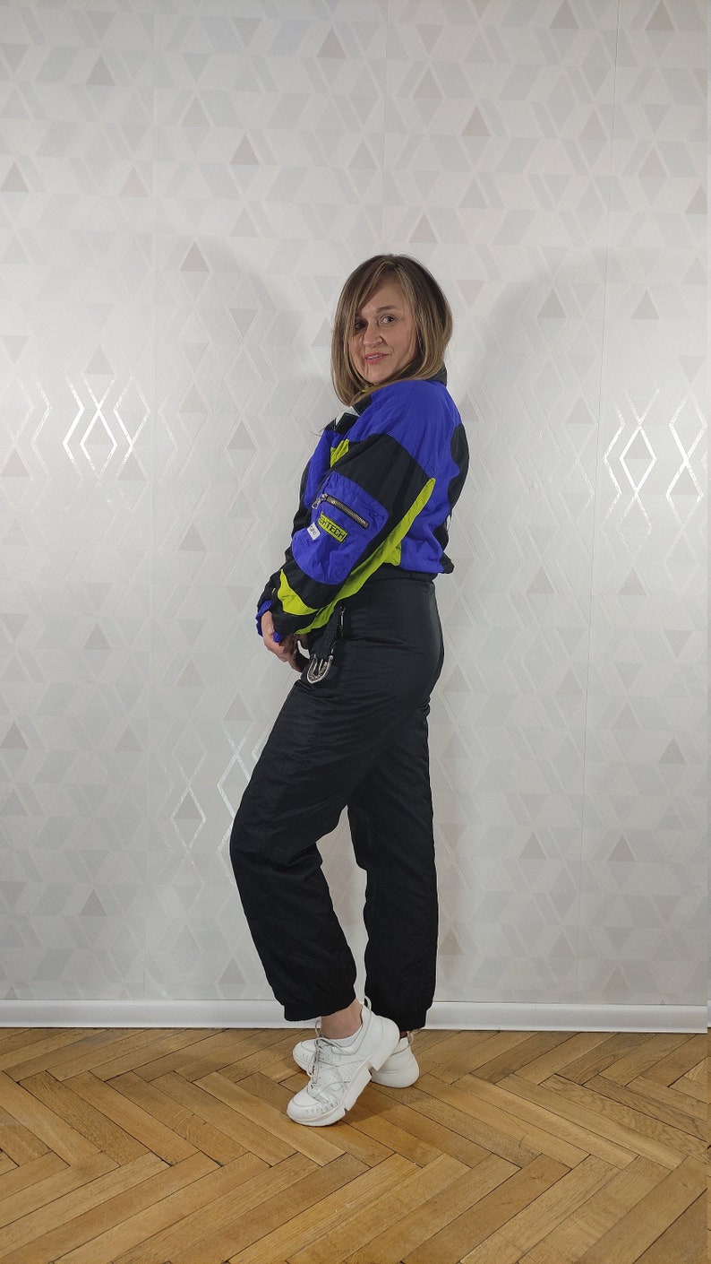 Pode incluir: Um macac&atilde;o de esqui preto com uma jaqueta de esqui azul e verde neon. A jaqueta tem um z&iacute;per e v&aacute;rios bolsos. O macac&atilde;o est&aacute; sendo usado por uma pessoa com t&ecirc;nis brancos.
