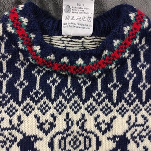 Puede incluir: Un su&eacute;ter de punto azul y blanco con un patr&oacute;n tradicional noruego. El su&eacute;ter tiene un ribete rojo y verde alrededor del cuello. La etiqueta en el su&eacute;ter dice "SIZE: L PURE NEW WOOL PURE LAINE REINE SCHURWOLLE JALE GARN & TRIKOTAS JE A/S 5280 DALEKVAM-NORWAY".