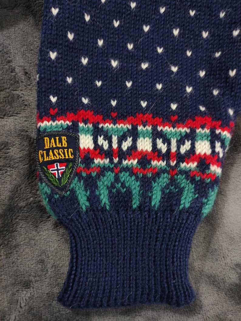 Puede incluir: Un su&eacute;ter de punto azul con un patr&oacute;n de corazones blancos y una banda con patr&oacute;n rojo, blanco y verde. El su&eacute;ter tiene una etiqueta que dice "DALE CLASSIC" con una bandera noruega.