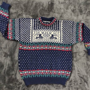 Puede incluir: Un su&eacute;ter de punto azul con un patr&oacute;n geom&eacute;trico blanco, rojo y verde. El su&eacute;ter tiene cuello redondo y mangas largas.