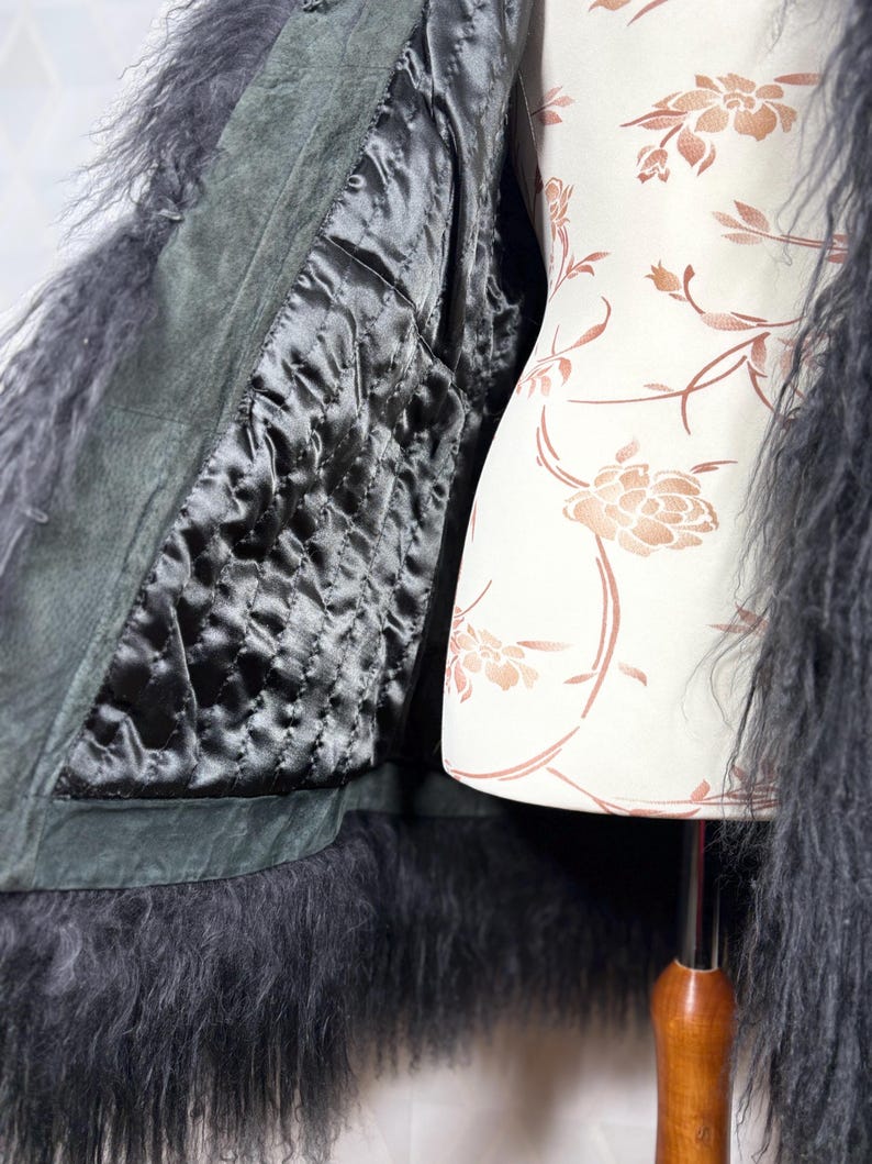 May include: A black faux fur jacket with a dark green suede lining. The jacket is open and the lining is visible. The jacket is on a white mannequin with a floral pattern.