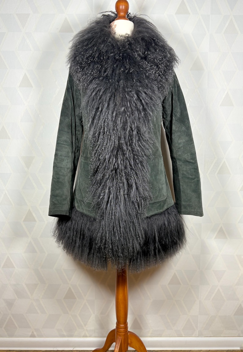 May include: A long, dark green suede coat with a large, fluffy gray fur collar and trim. The coat has a long, flowing silhouette and a relaxed fit.