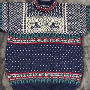 Puede incluir: Un su&eacute;ter de punto azul, blanco, rojo y verde con cuello redondo. El su&eacute;ter tiene un dise&ntilde;o geom&eacute;trico con un patr&oacute;n de reno en el centro.