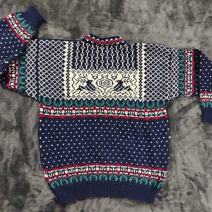 Puede incluir: Un su&eacute;ter de punto azul con un dise&ntilde;o estampado en blanco, rojo y verde. El su&eacute;ter presenta una secci&oacute;n blanca con un dise&ntilde;o estilizado de dos p&aacute;jaros y un ciervo.