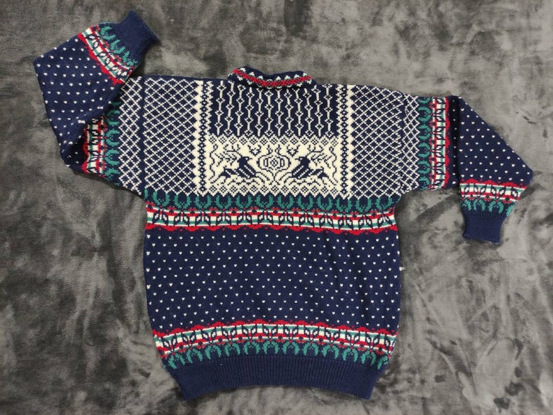 Puede incluir: Un su&eacute;ter de punto azul con un dise&ntilde;o estampado en blanco, rojo y verde. El su&eacute;ter presenta una secci&oacute;n blanca con un dise&ntilde;o estilizado de dos p&aacute;jaros y un ciervo.