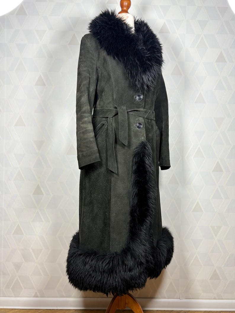 Puede incluir: Abrigo largo de ante verde oscuro con cuello, pu&ntilde;os y ribetes de piel negra. El abrigo tiene cintur&oacute;n en la cintura y cierres de botones. El abrigo vintage se exhibe en un maniqu&iacute;.