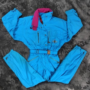 Op de afbeelding: Een felblauwe skibroek met een roze kraag en een zwart logo met de tekst "Snow Tech". De broek heeft een riem en meerdere zakken.