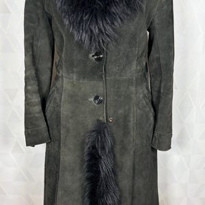Puede incluir: Un abrigo de ante verde oscuro con ribete de piel sint&eacute;tica negra alrededor del cuello, en la parte delantera y en el dobladillo. El abrigo tiene cierre de botones y mangas largas. El abrigo es de estilo largo y elegante.