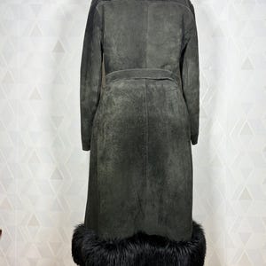 Puede incluir: Un abrigo largo de ante gris oscuro con cuello y ribete de piel negra. El abrigo tiene cintur&oacute;n en la cintura y se exhibe sobre un soporte de madera. El dise&ntilde;o del abrigo es vintage y elegante.
