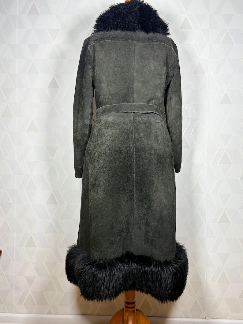 Puede incluir: Un abrigo largo de ante gris oscuro con cuello y ribete de piel negra. El abrigo tiene cintur&oacute;n en la cintura y se exhibe sobre un soporte de madera. El dise&ntilde;o del abrigo es vintage y elegante.