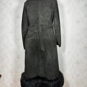 Puede incluir: Un abrigo de ante gris oscuro con cuello y ribete de piel negra. El abrigo largo presenta un cintur&oacute;n a juego y se exhibe sobre un soporte de madera. La vista trasera muestra el dise&ntilde;o y la textura del abrigo.
