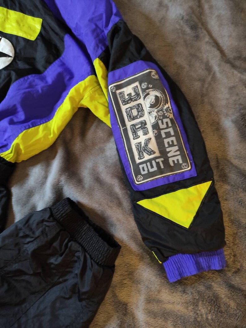 Pode incluir: Casaco preto, roxo e amarelo neon com um remendo preto na manga que diz "OUT SCENE 30K".