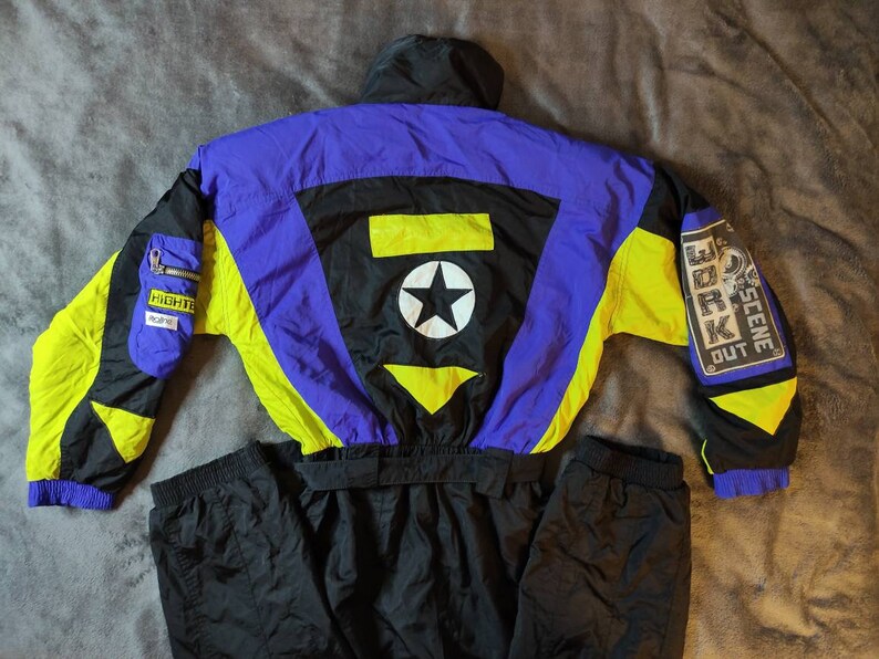 Pode incluir: Um macac&atilde;o de esqui preto, amarelo e azul com uma estrela e as palavras "SCENE OUT WORK" nas costas.