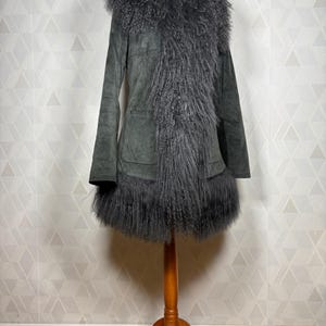 May include: A gray suede coat with a long, fluffy gray fur collar and trim. The coat has a long, fitted silhouette and a single pocket on the front.