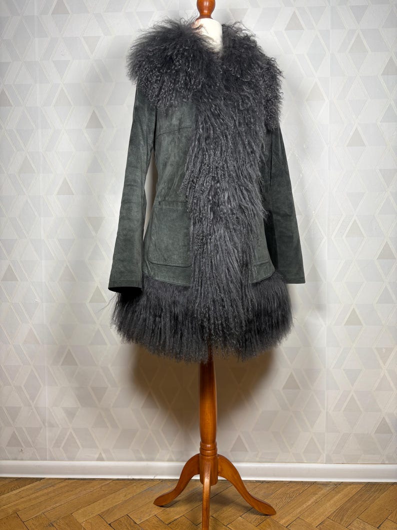 May include: A gray suede coat with a long, fluffy gray fur collar and trim. The coat has a long, fitted silhouette and a single pocket on the front.