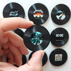 May include: A collection of black vinyl record magnets featuring album art and band names like Daft Punk, Metallica, and The Beatles. One magnet is held in a hand, with others arranged on a white surface.