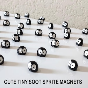 May include: A collection of black and white soot sprite magnets. The magnets are small and round, with two white eyes on each. The magnets are arranged in rows on a white surface. The text "CUTE TINY SOOT SPRITE MAGNETS" is printed below the magnets.